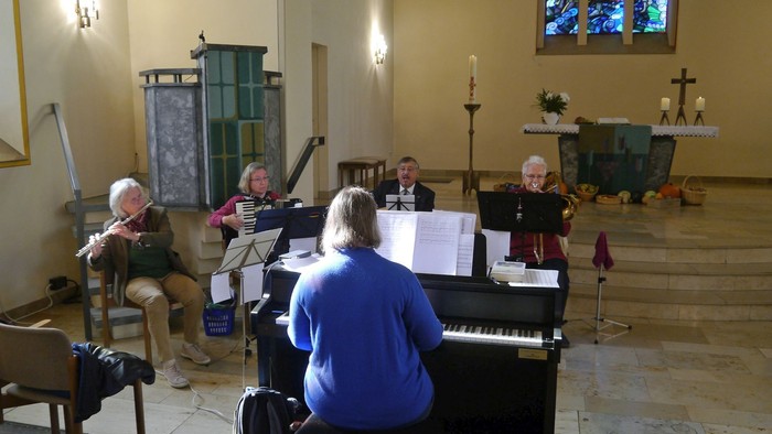 Band spielt bei einem Auftritt in der Kreuzkirche; zu sehen sind die Querflötistin, die Akkordeonspielerin, der Gitarrist und die Posaunistin, jeweils mit ihrem Instrument; im Vordergrund (mit dem Rücken zur Kamera) die Leiterin am E-Piano